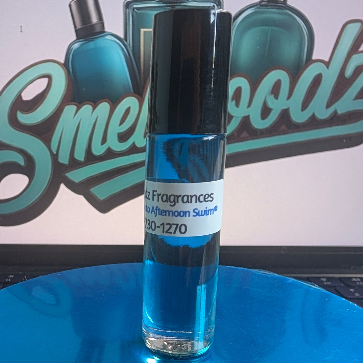 A small glass roll-on bottle with blue liquid and a black cap stands on a reflective blue circular platform. The bottle label reads 'Fragrances'